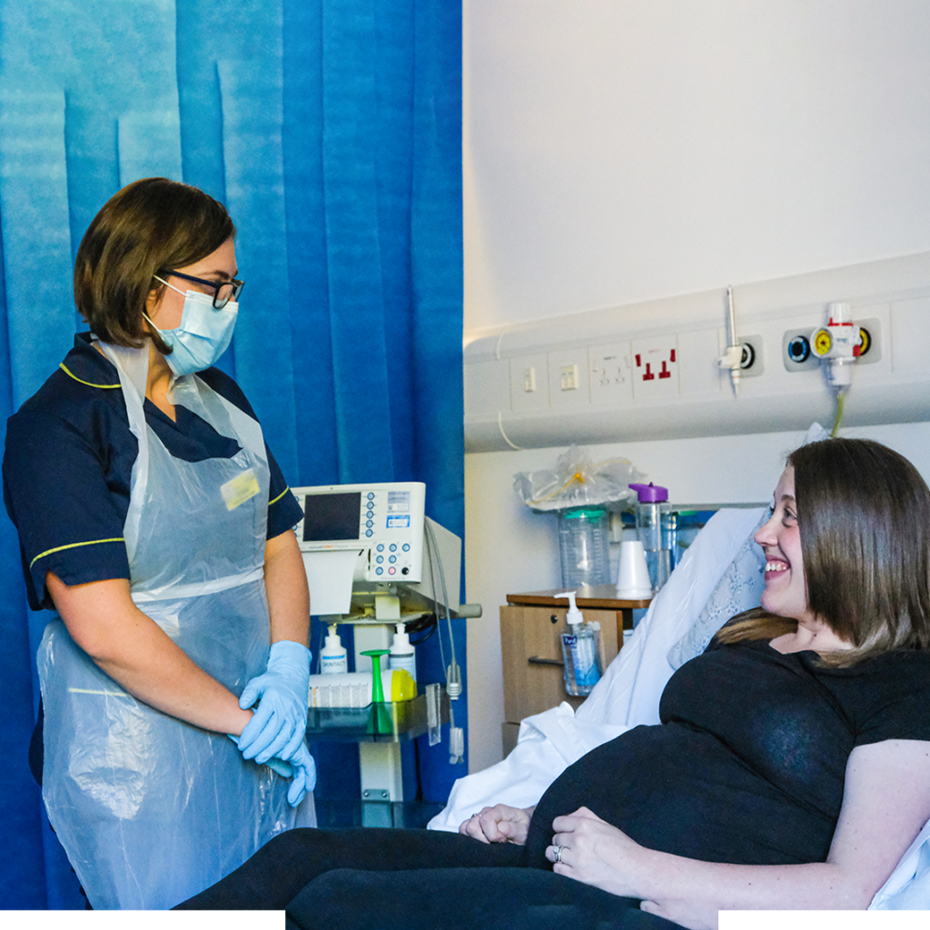 Mum discussing with the nurse A midwife dressed in PPE standing in a clinical bay looks down at a smiling pregnant woman who is dressed casually and laying on a hospital bed.
